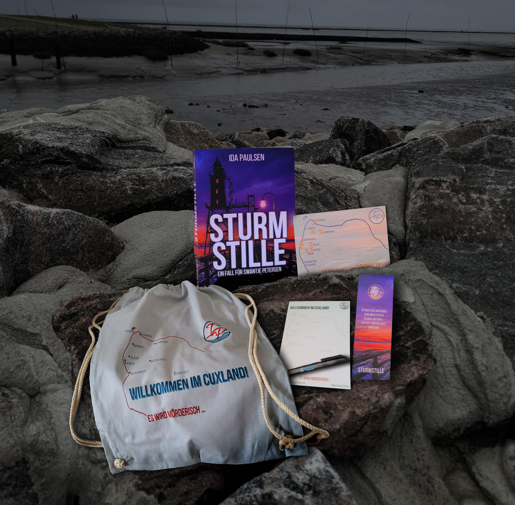 Premiumbundle zu Sturmstille Auf dem Bild sieht man die einzelnen Elemente des Premiumbundles (Taschenbuch Sturmstille, Turnbeutel, Block und Kugelschreiber, Lesezeichen und Postkarte), die auf einer steinernen Begrenzung zum Wattenmeer liegen.